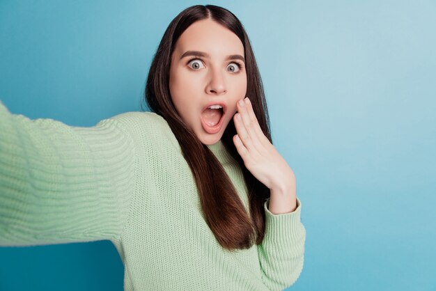 Portrait of shocked adorable girl hold camera take selfie open mouth on teal background