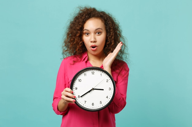 Portrait of shocked amazed african girl in pink casual clothes holding round clock isolated on blue turquoise wall background in studio. People sincere emotions, lifestyle concept. Mock up copy space.