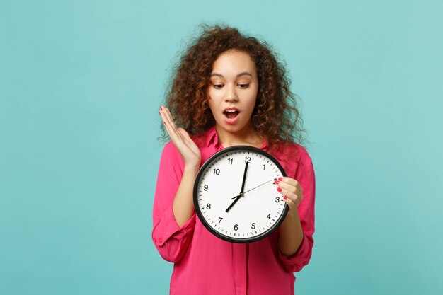 Portrait of shocked amazed african girl in pink casual clothes holding round clock isolated on blue turquoise wall background in studio. People sincere emotions, lifestyle concept. Mock up copy space.