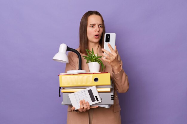 Portrait of shocked amazed Caucasian woman wearing official style jacket holding folders isolated over purple background using cell phone reading message with astonished information