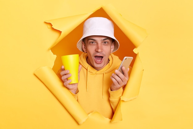 Portrait of shocked amazed man wearing hoodie and panama looking at camera with open mouth and holding cup using cell phone for browsing Internet breaking through torn in yellow paper wall