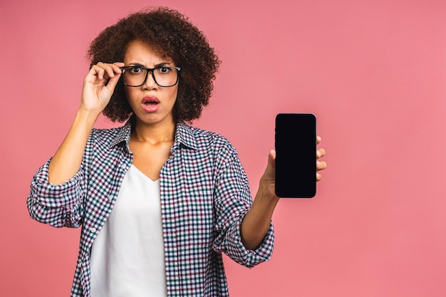 Portrait of a shocked amazed surprised young african american\
woman holding blank screen mobile phone isolated over pink\
background