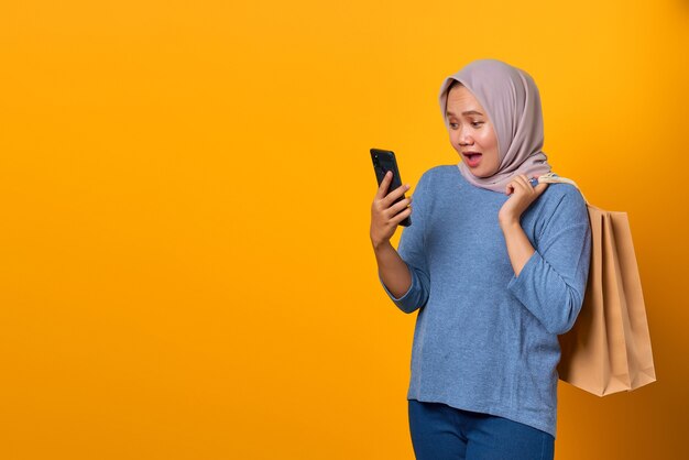 Portrait of shocked Asian woman holding phone and shopping bag over yellow background