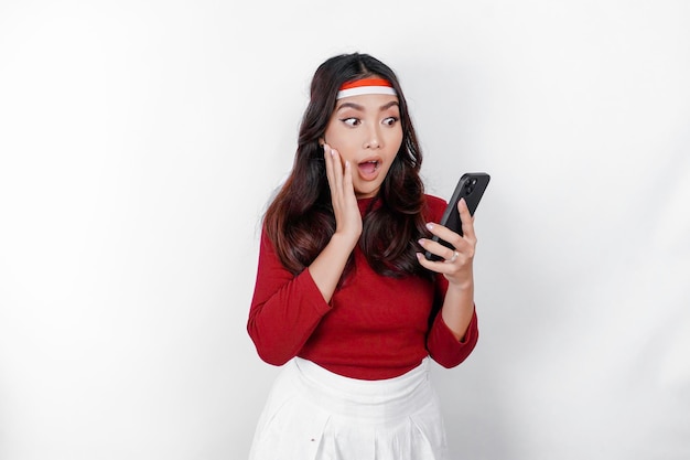 A portrait of a shocked Asian woman wearing headband holding her phone while her mouth wide open isolated by white background Indonesia39s independence day concept