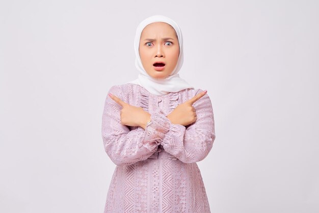 Portrait of shocked beautiful Asian Muslim woman wearing hijab and purple dress Pointing index fingers aside isolated over white background