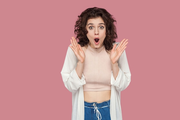 Portrait of shocked beautiful brunette young woman with curly hairstyle in casual style standing with raised arms and looking at camera with amazed face. indoor studio shot isolated on pink background