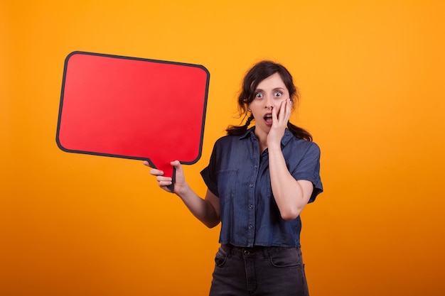 Portrait of shocked beautiful woman holding empty think bubble in studio over yellow background. Red bubble thoughts space.