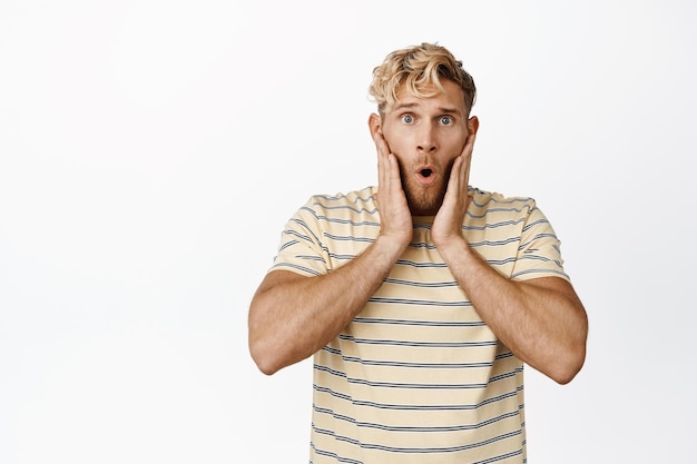 Portrait of shocked blond guy holding hands on face saying wow and staring impressed at camera standing over white background