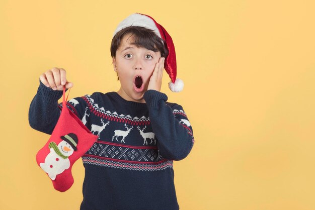 Portrait of shocked boy wearing santa hat holding christmas decoration against yellow background