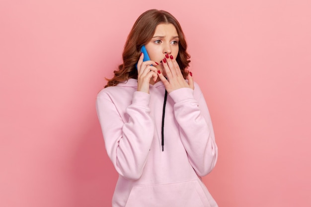 Portrait of shocked brunette teenage girl in casual hoodie talking smartphone and covering mouth with palm bad news communication Indoor studio shot isolated on pink background