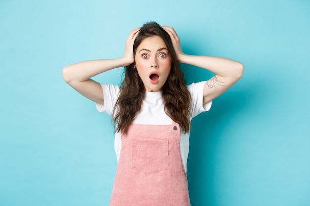 Portrait of shocked brunette woman holding hands on head and gasping, staring at camera troubled, having big problem, standing against blue background