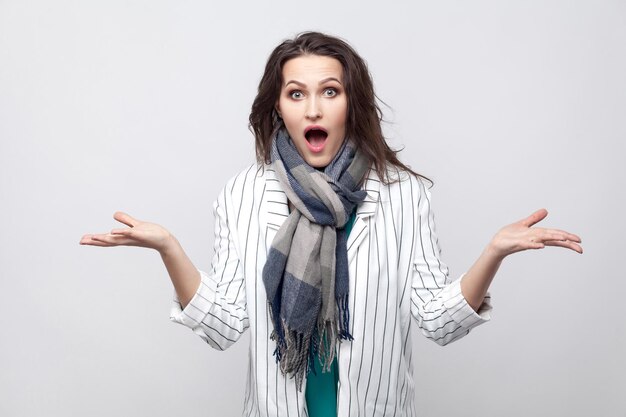 Portrait of shocked brunette woman in white striped jacket and blue scarf standing, looking at camera with big eyes and raised arms and screaming. indoor studio shot, isolated on light grey background