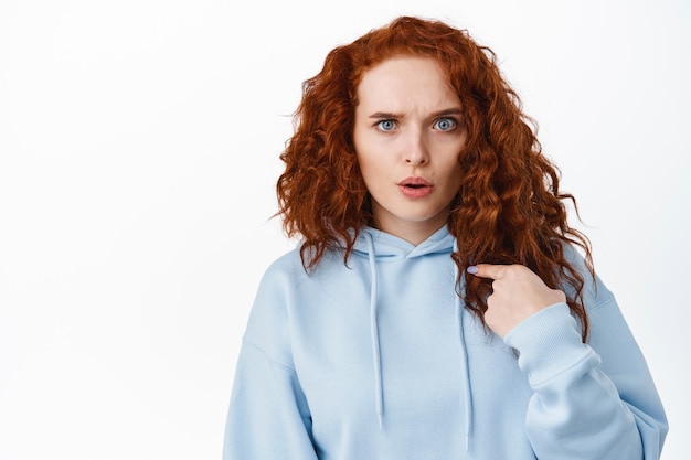 Portrait of shocked and confused ginger girl pointing at herself, being accused or blamed in something, stare  questioned, white wall