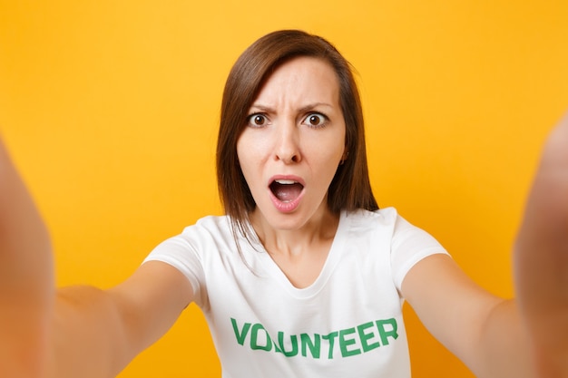 Portrait of shocked displeased irritated woman in white t-shirt with written inscription green title volunteer isolated on yellow background. Voluntary free assistance help, charity grace work concept