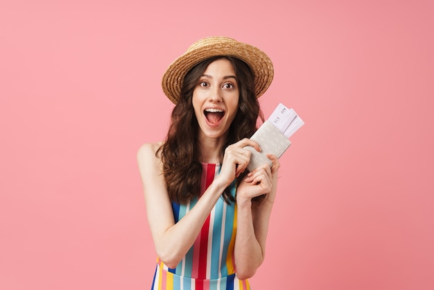 Portrait of shocked emotional happy young cute woman posing isolated over pink wall holding passport with tickets