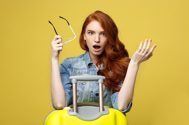 Portrait of a shocked girl in denim dress with suitcase looking at camera.