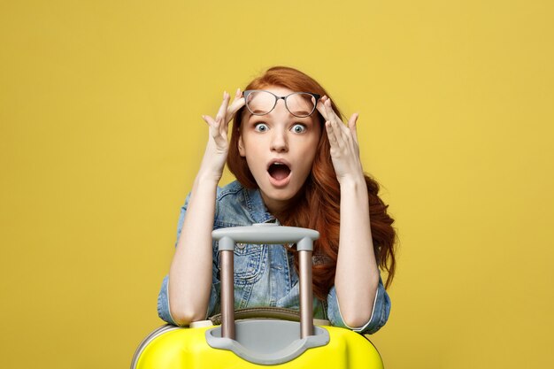 Portrait of a shocked girl in denim dress with suitcase looking at camera.