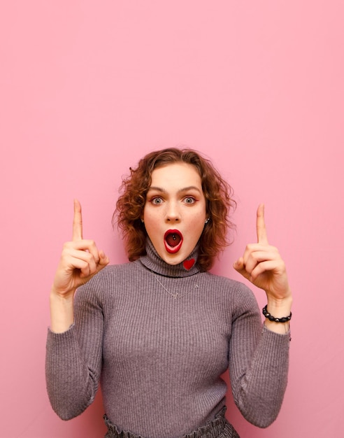 Portrait of shocked girl in gray sweater and with curly hair on pink background looks into camera