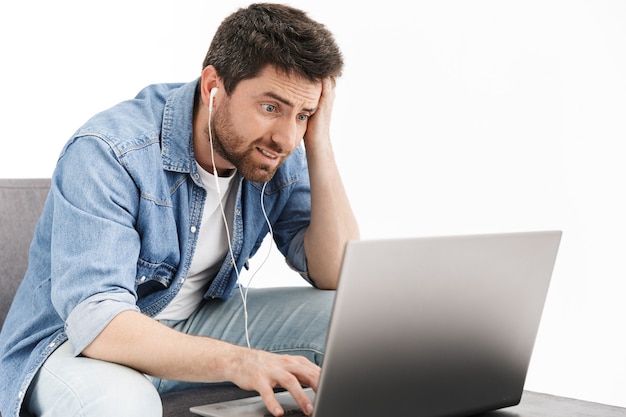 Portrait of a shocked handsome bearded man wearing casual clothes sitting in chair isolated, working on laptop computer