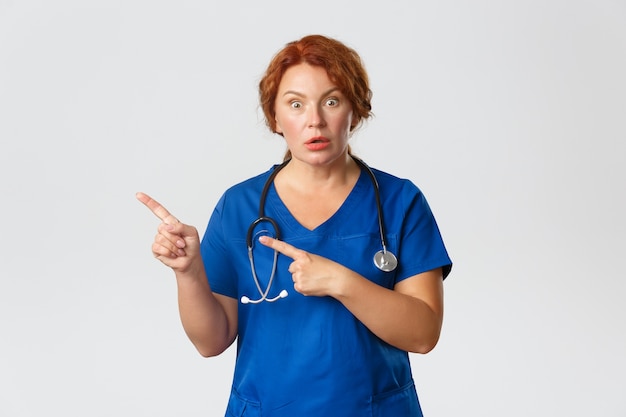 Portrait of shocked and impressed redhead female doctor, nurse in scrubs pointing fingers left on grey