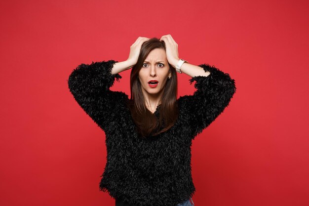 Portrait of shocked perplexed young woman in black fur sweater clinging to head isolated on bright red wall background in studio. People sincere emotions, lifestyle concept. Mock up copy space.