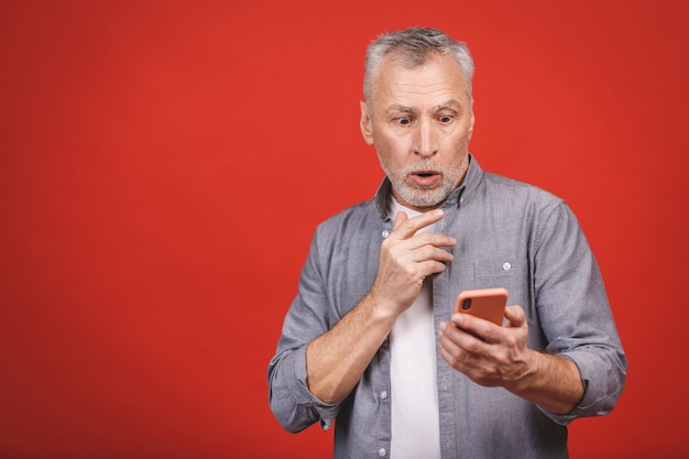 Portrait of shocked senior man in casual gasping opening mouth from worry and surprise holding mobile phone, looking in camera isolated over red wall.