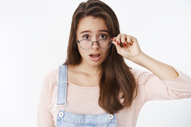 Portrait of shocked speechless and questioned female brunette taking off glasses dropping jaw and raising eyebrows from amazement and stupod over gray wall