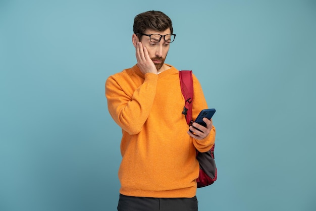 Portrait of shocked student man reading something using cell phone looking surprised Indoor studio shot isolated on blue background