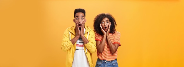 Portrait of shocked and stunned speechless girlfriend dropping jaw from amazement with boyfriend feeling amazed from shook news posing together surprised and astonished over orange wall