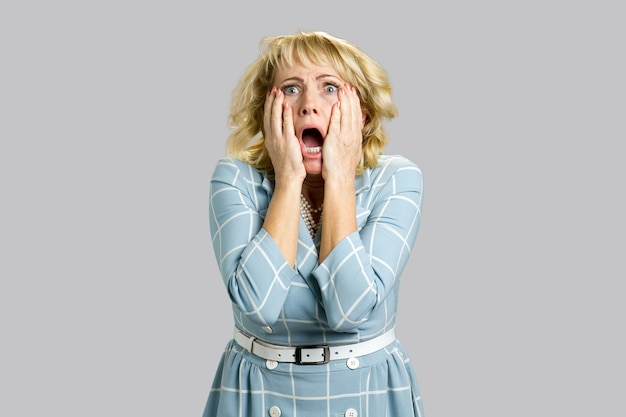 Portrait of shocked and stunned woman. Mature woman looking excited in full disbelief, open mouth and hands on face, grey. Negative human face expression.