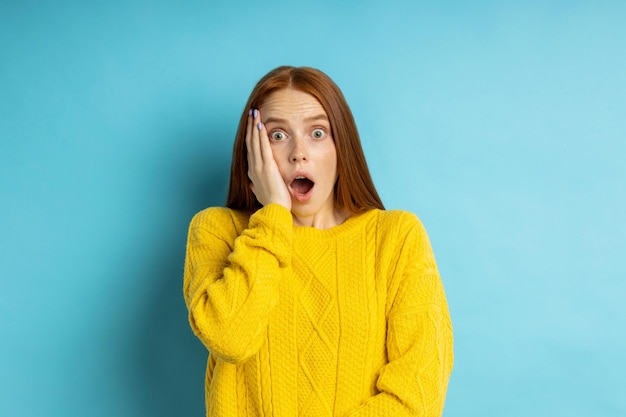 Portrait of shocked stunned young ginger female looking at camera with opened mouth, keeping hands on cheeks, standing against blue background. Expressive facial expressions, reaction concept.