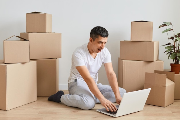 Portrait of shocked surprised man wearing casual clothing sitting on floor near carton parcels and typing on notebook keyboard expressing fear and astonishment forgot about important task