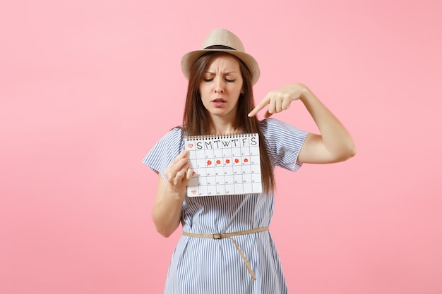 Portrait of shocked woman in blue dress, hat holding periods calendar for checking menstruation days isolated on bright trending pink background. Medical, healthcare, gynecological concept. Copy space