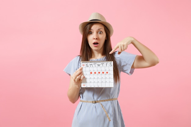 Ritratto di donna scioccata in abito blu, cappello che tiene il calendario dei periodi per controllare i giorni delle mestruazioni isolati su sfondo rosa di tendenza brillante. concetto medico, sanitario, ginecologico. copia spazio