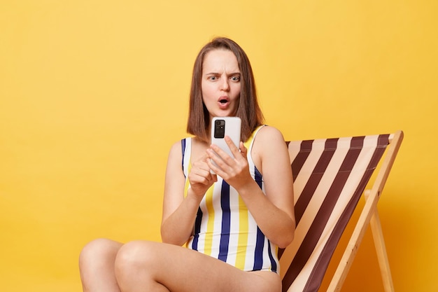 Portrait of shocked woman sits on deck chair holding mobile phone communicates in online chat against yellow background reading shocked or bad news checking free dates for departure has bad result