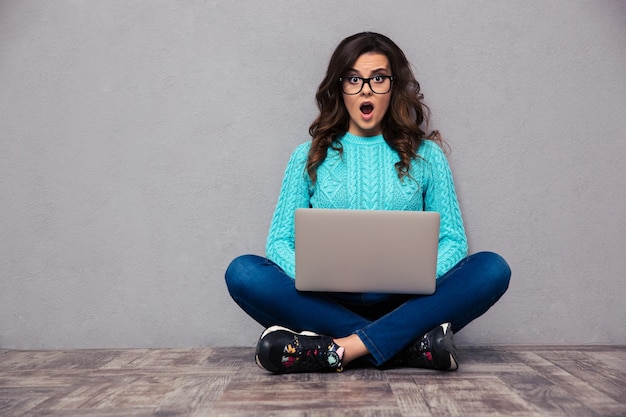 Portrait of shocked woman sitting on the floor with laptop and  on gray wall