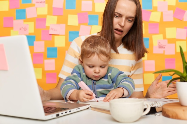 Photo portrait of shocked woman with brown hair wearing striped tshirt sitting at table standing against yellow wall with colorful sticky notes mother with kid in office child messes up mommy's documents