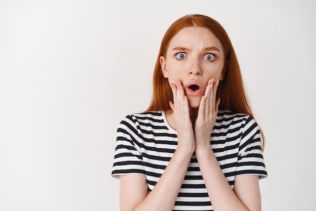 Portrait of shocked and worried redhead woman hear bad news, holding hands on face and frownign, staring at at front startled, white wall