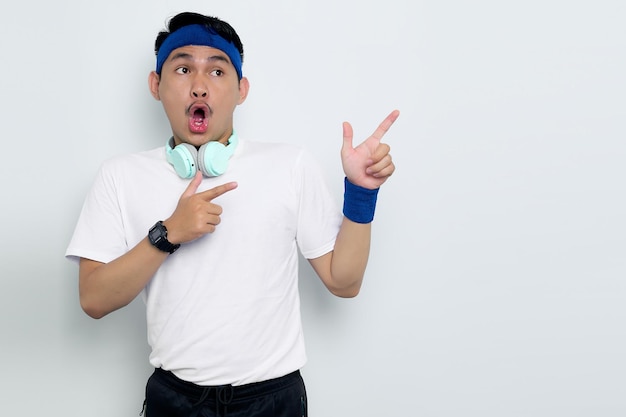 Портрет шокированного молодого азиатского спортсмена в синей повязке на голове и спортивной белой футболке с наушниками, указывающими пальцами на рекламный баннер копировального пространства на белом фоне