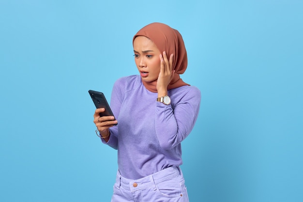 Портрет потрясенной молодой азиатской женщины, использующей мобильный телефон на синем фоне