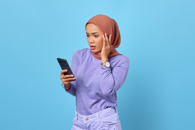 青い背景の上の携帯電話を使用してショックを受けた若いアジアの女性の肖像画