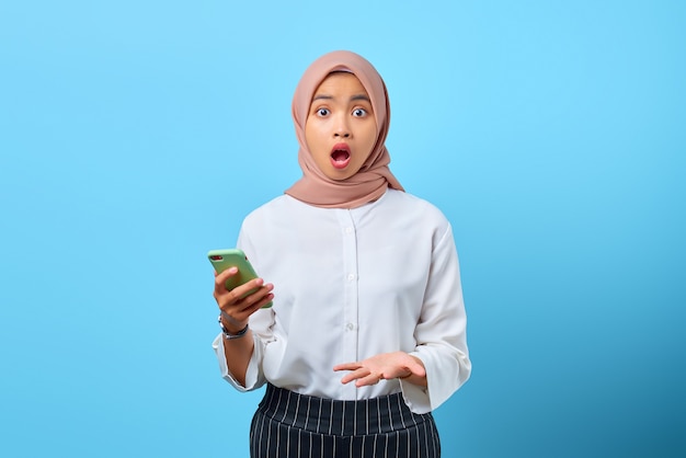 Portrait of shocked young Asian woman using mobile phone with open mouth over blue background