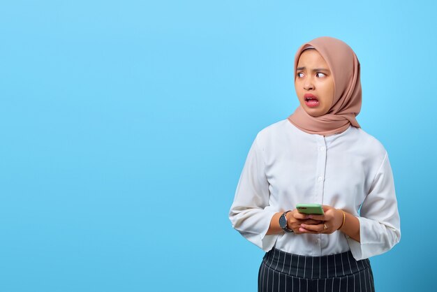 Portrait of shocked young Asian woman using mobile phone with open mouth and looking to sideways