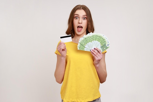 Portrait of shocked young female holding euro banknotes and credit card unbelievable bank loan cashback looking at camera with opened mouth Indoor studio shot isolated on gray background