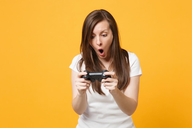 Portrait of shocked young girl in white casual clothes keeping mouth open playing video game with joystick isolated on yellow orange background in studio. People lifestyle concept. Mock up copy space.