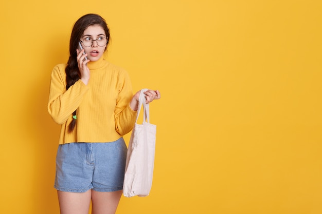 Portrait of shocked young girl with long dark hair posing isolated over yellow wall with phone, talking to somebody with astonished facial expression