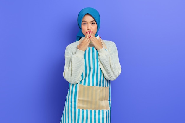 히잡과 앞치마를 입은 충격을 받은 젊은 주부 여성의 초상화는 보라색 배경에 격리된 손으로 입을 가리고 있는 카메라를 보고 있는 주부 이슬람 생활 방식 개념입니다.