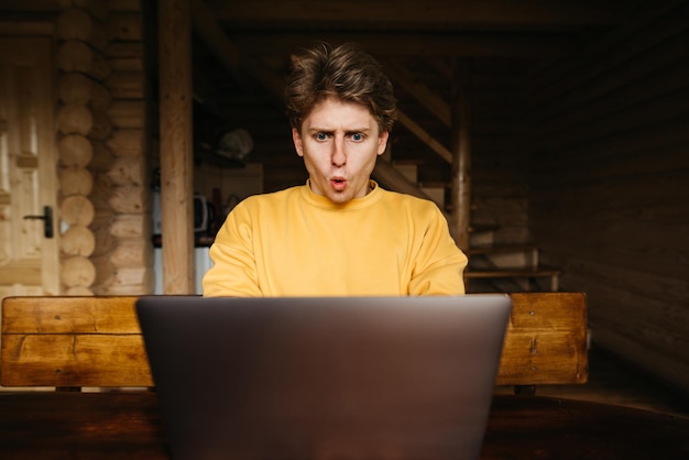 Portrait of shocked young man in casual clothes using laptop in wooden country house