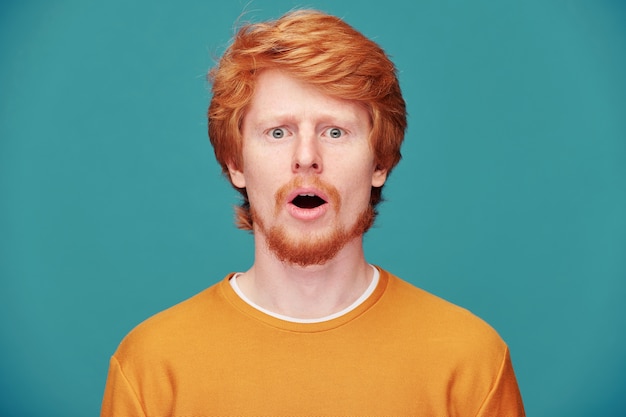 Portrait of shocked young redhead guy with beard keeping mouth open in astonishment on blue