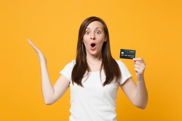 Portrait of shocked young woman in casual clothes keeping mouth open, spreading hands, holding credit bank card isolated on yellow orange wall background. People lifestyle concept. Mock up copy space.